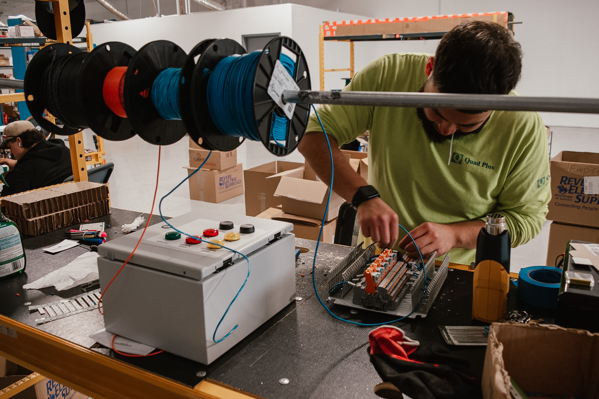 Close-up of a technician wiring an electrical control panel, connecting blue wires to terminal blocks inside a cable management tray during assembly.
